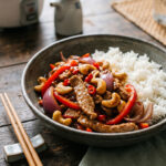 Eine rustikale Schale mit asiatisch gebratenem Schweinefilet in feinen Streifen, roten Paprikastreifen, Zwiebeln und gerösteten Cashewkernen in einer glänzenden Sauce, serviert mit fluffigem weißem Reis und Stäbchen auf einem Holztisch.