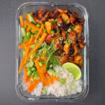 Gochujang-Hähnchen mit glänzender Marinade, kombiniert mit einem knackigen Edamame-Salat und feinen Gemüsestiften. Die frische Minze und die Limette setzen farbliche Akzente im Glascontainer auf puristischem grauem Grund.